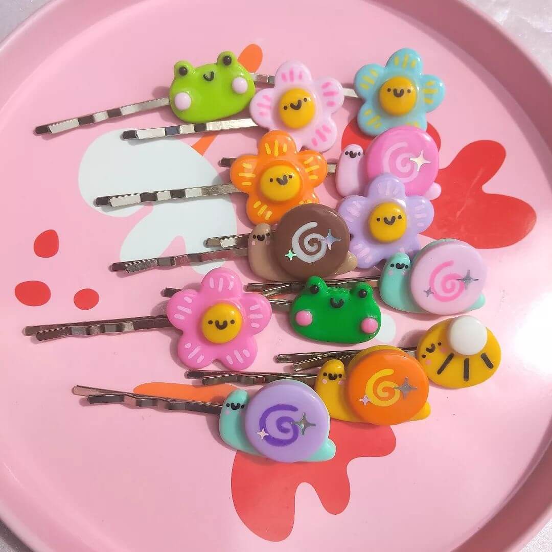 Colourful hair clips made from polymer clay with cartoon frogs, flowers with faces, snails and bumble bees on a pink dish.