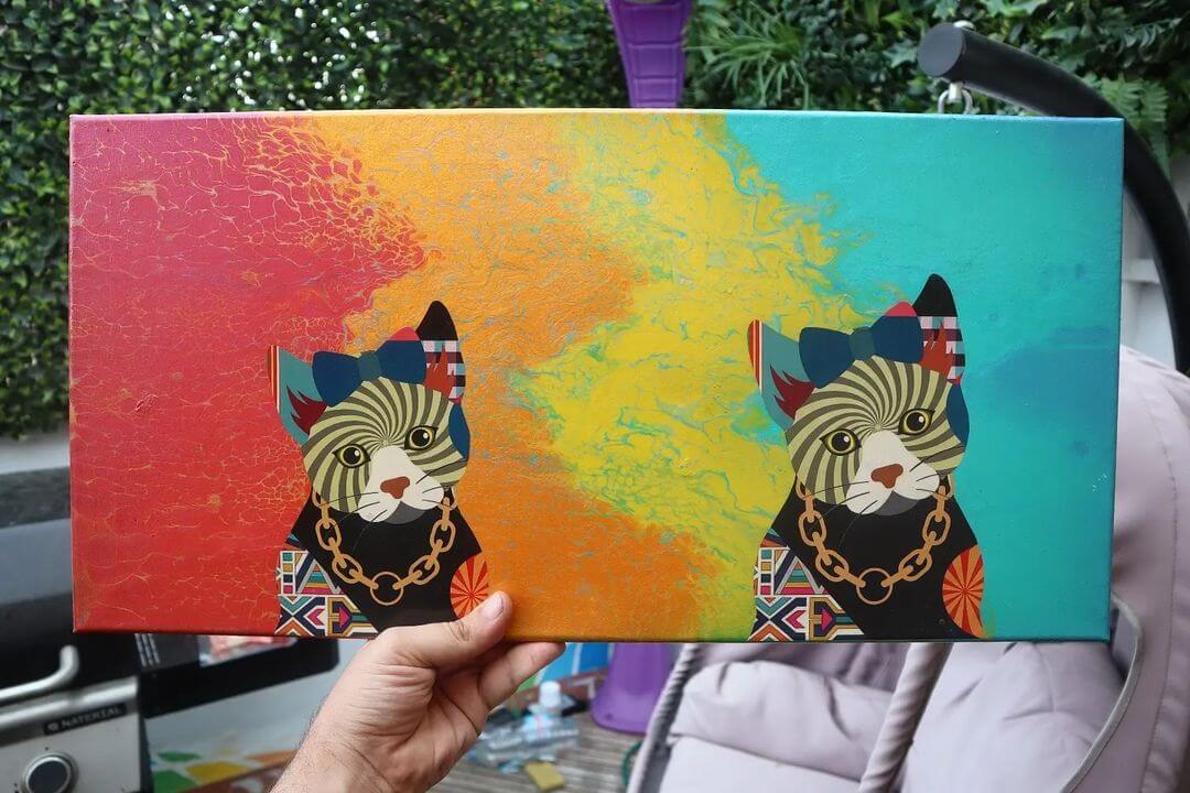 8. Hand holding a rainbow canvas with two cats painted on top wearing gold chains.