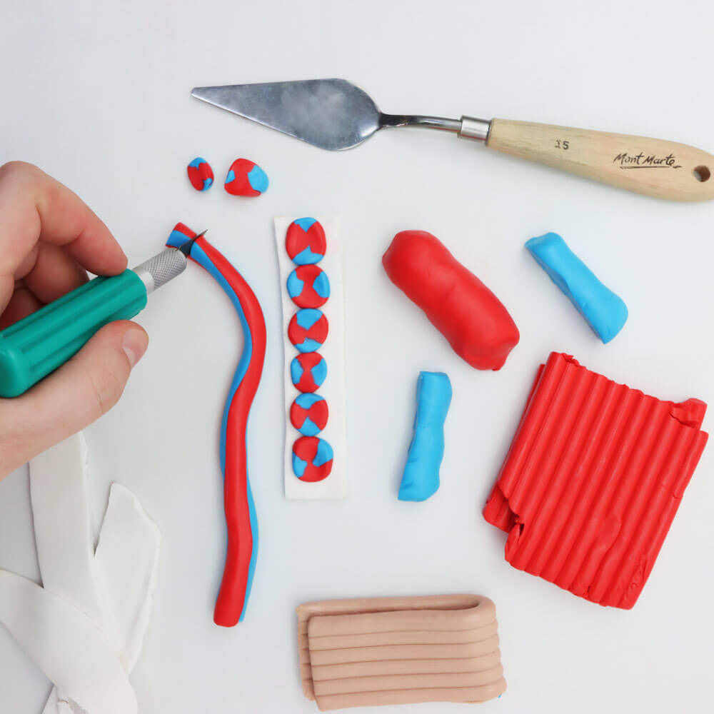 Hand holds a hobby knife to cut red and blue polymer clay. Red and blue clay blocks lay next to a palette knife.