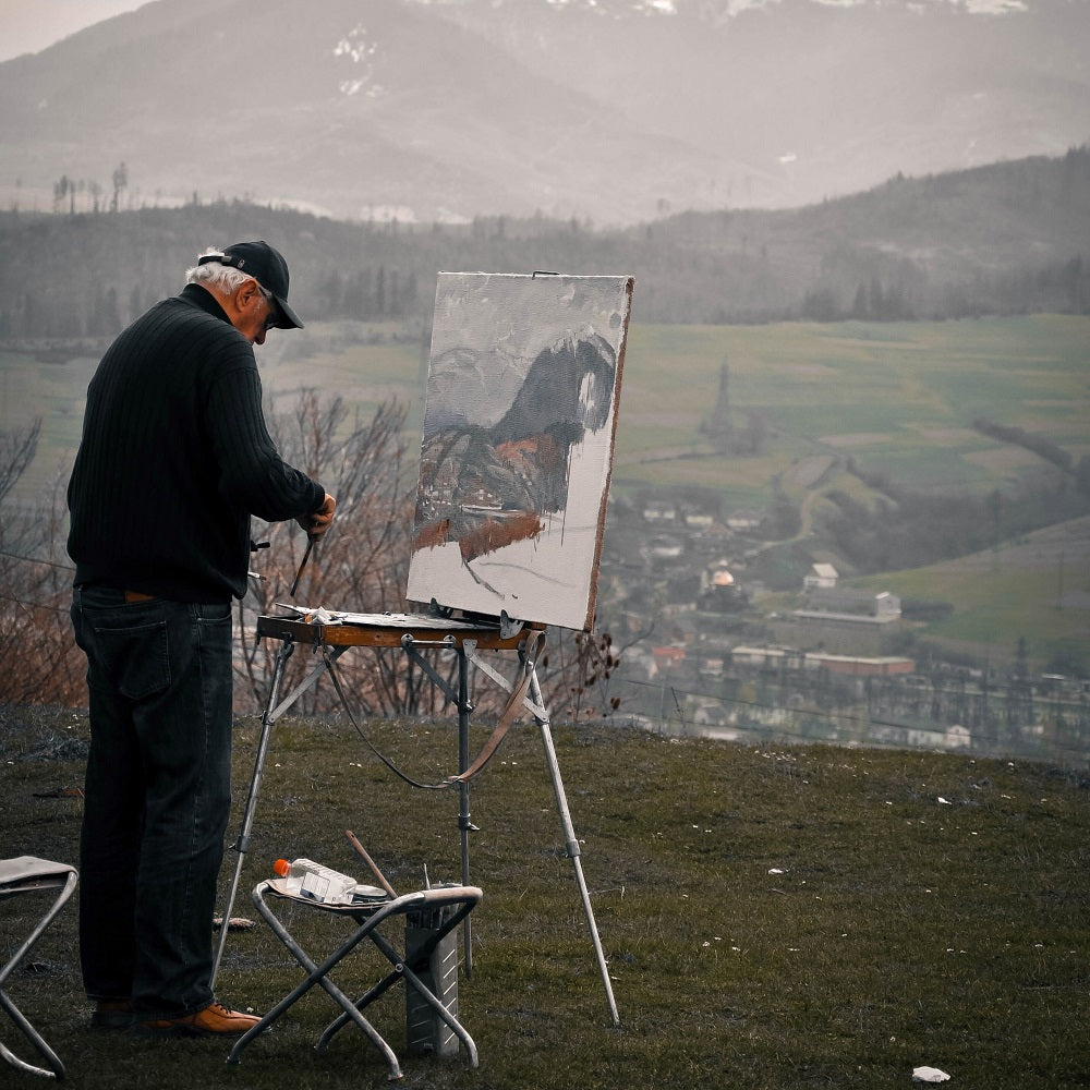 Man dressed in a jumper and jeans painting an outside landscape of a mountain on French easel.