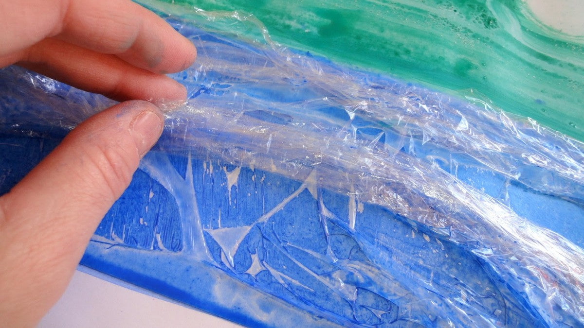 Hand placing cling film onto blue and green watercolour painting. 