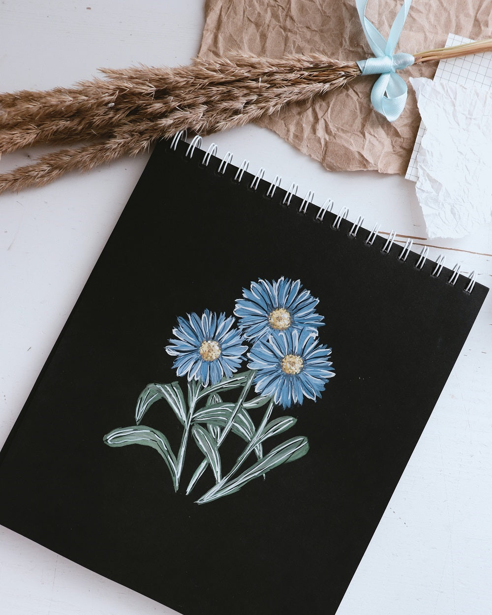 Three blue flowers drawn on a black paper pad in watercolour and gouache.