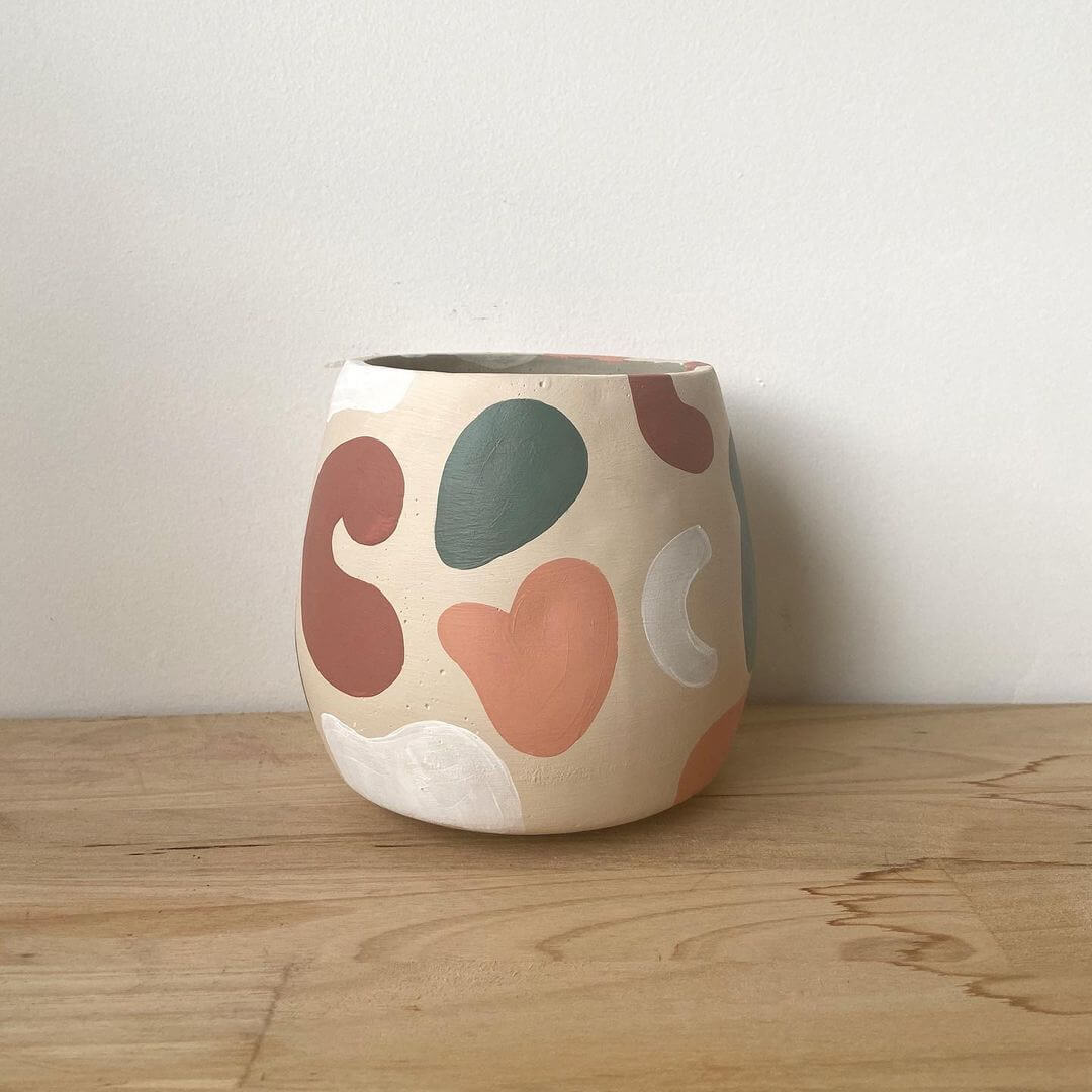 White pot with abstract shapes painted in earthy colours. Pot sits on a timber shelf next to a white wall.