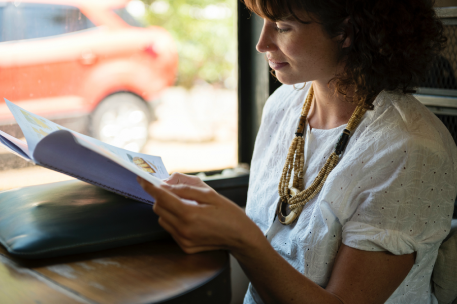 Female in a white blouse and beaded jewellery reading a magazine in a cafe near a window.