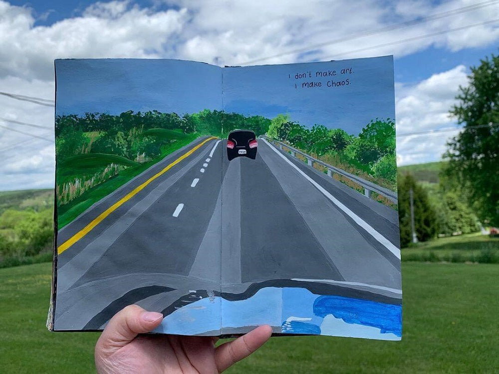 Hand holding a painting in a filed of a road with a car in front.