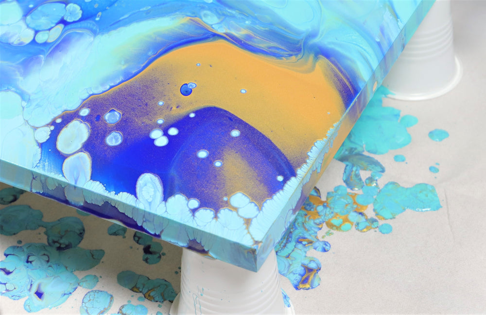 Teal, royal blue, and gold paints poured over the corner of a canvas being elevated by upside down cups
