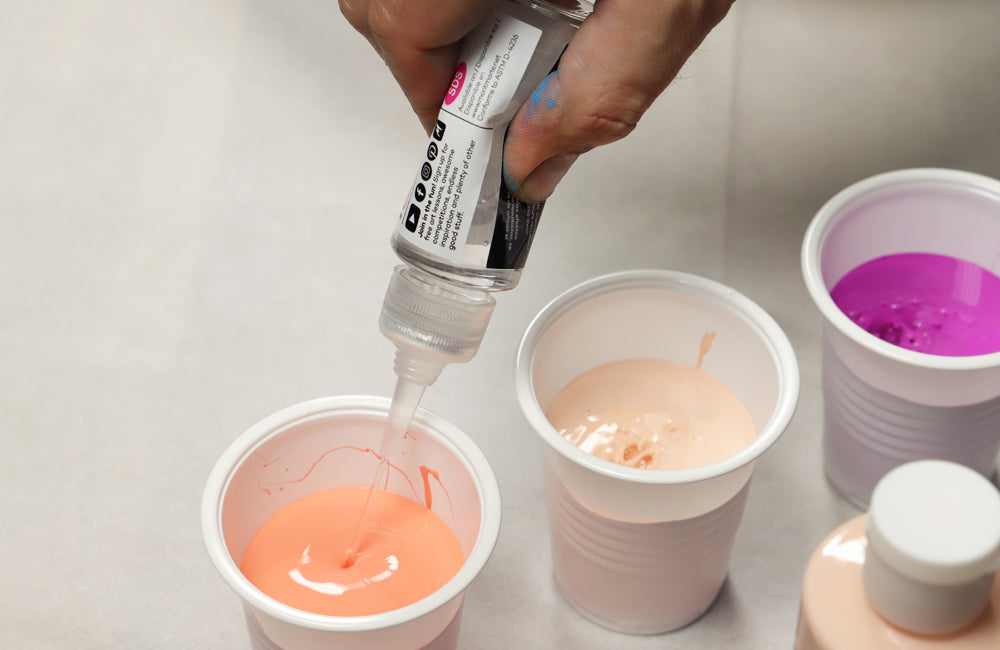 Silicone being added to a peach acrylic pouring paint in a white cup 