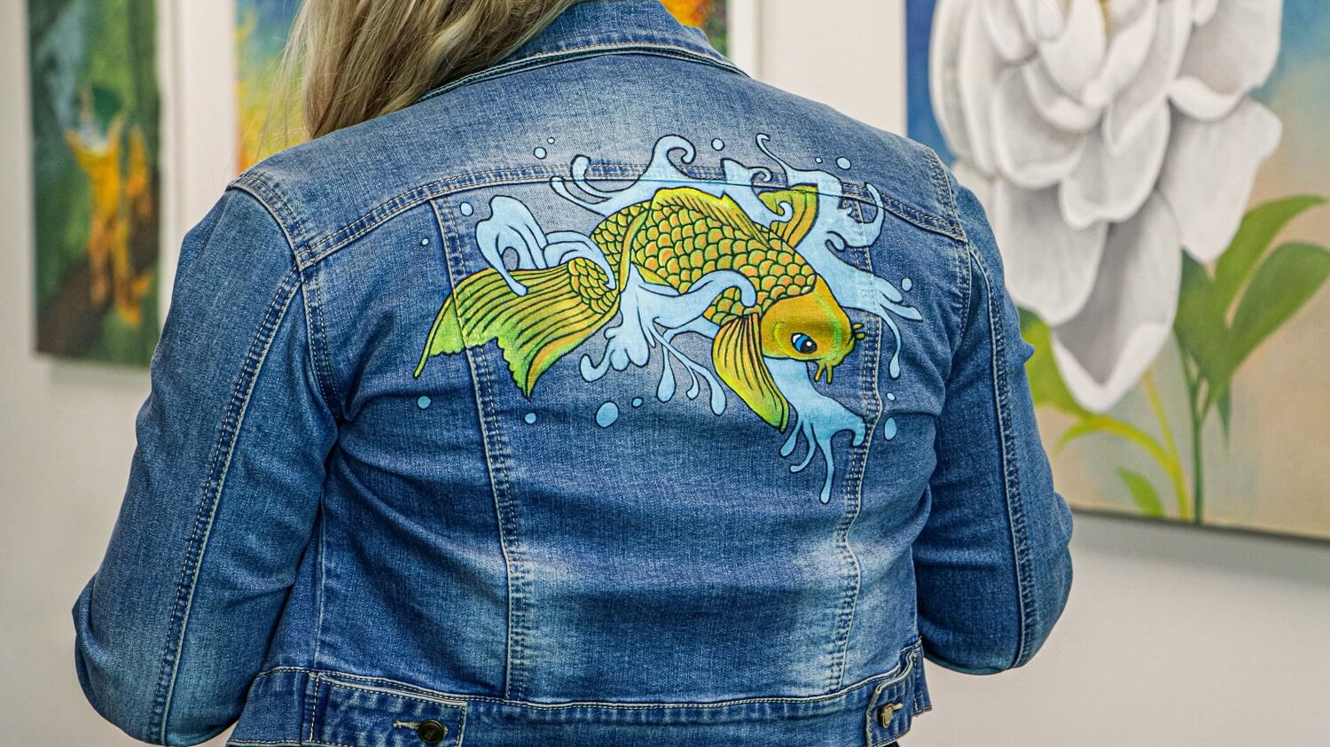 Blonde woman wearing a blue denim jacket with a yellow koi fish painted on the back.