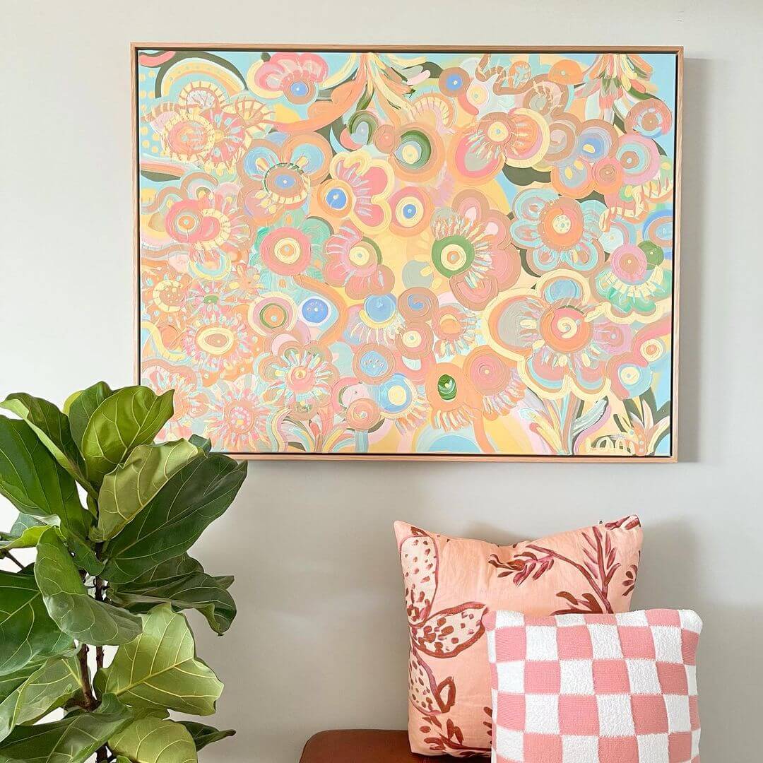 Colourful abstract artwork with pastel colours hanging on a wall next to two pink pillows and a green plant.