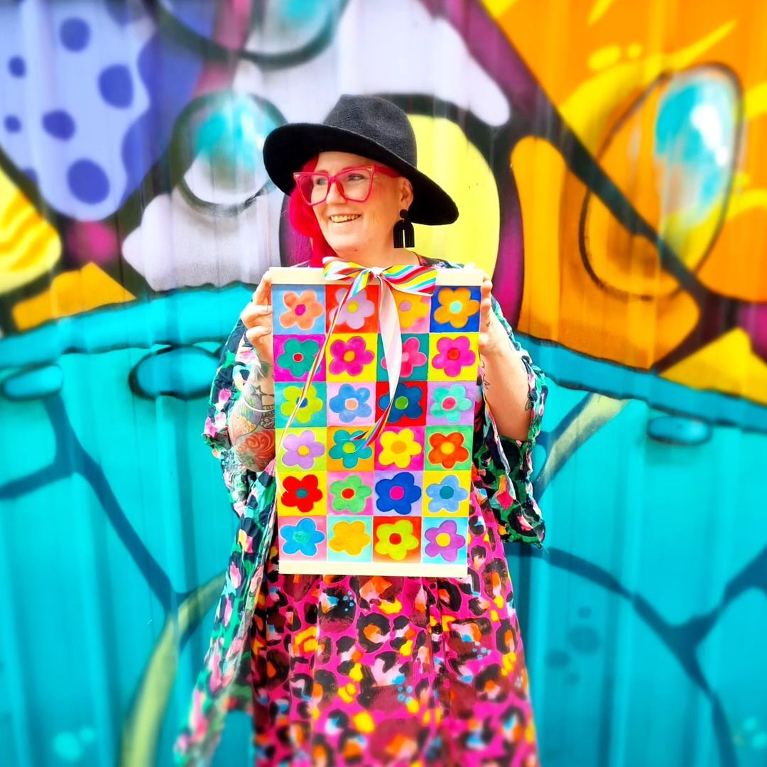 Ema Lou holding up a rainbow flower art painting against a colourful background