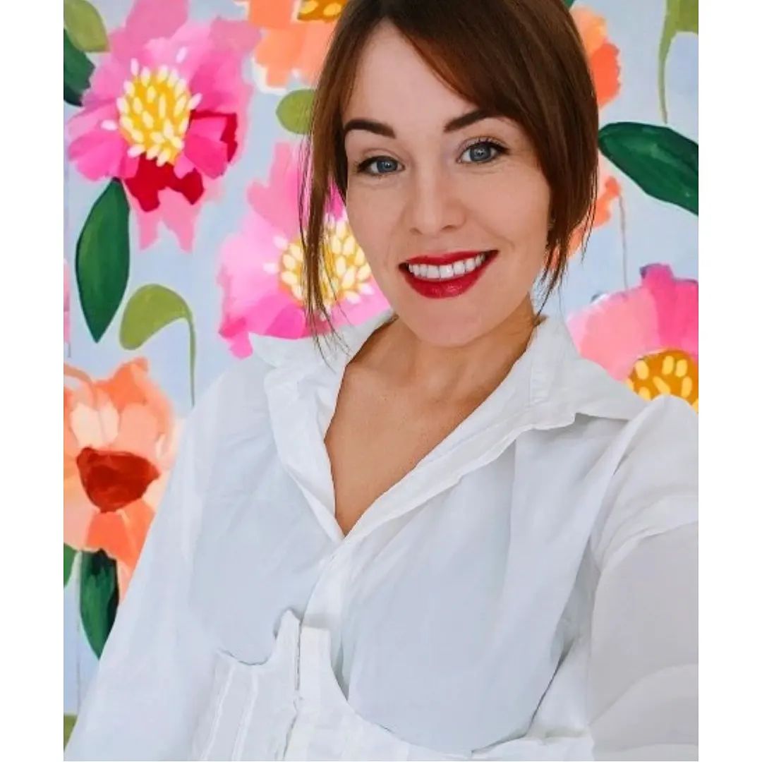 Emily Birks standing in front of one of her bright floral paintings in a white shirt