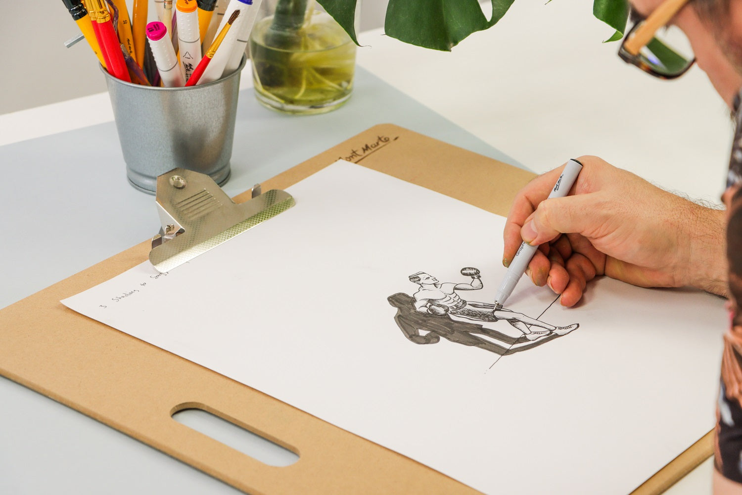 Man drawing a cartoon boxing character with a fine line marker next to a plant and a jar of markers.