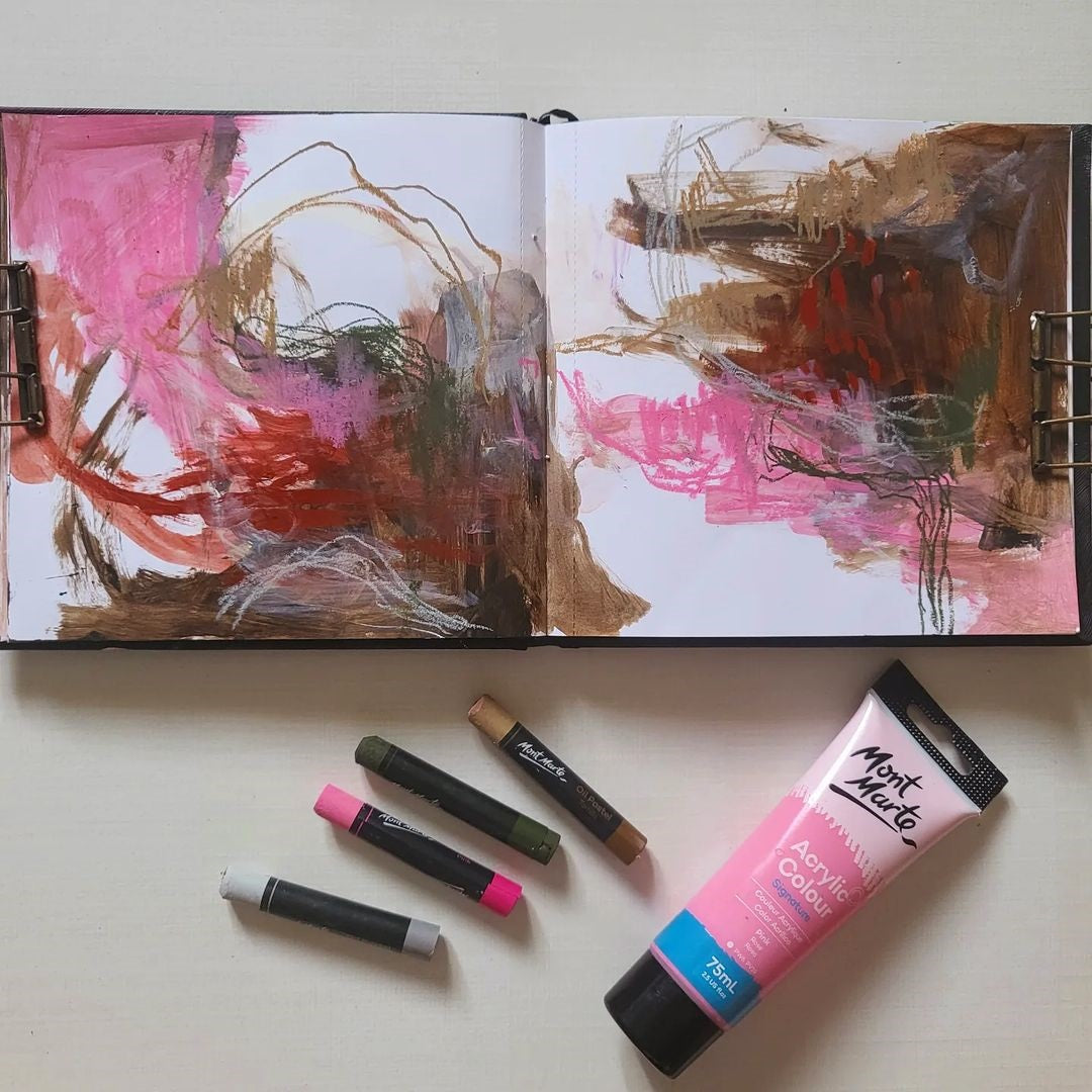 An open art journal with pink and earthy tones inside, next to four oil pastels and a pink paint tube.
