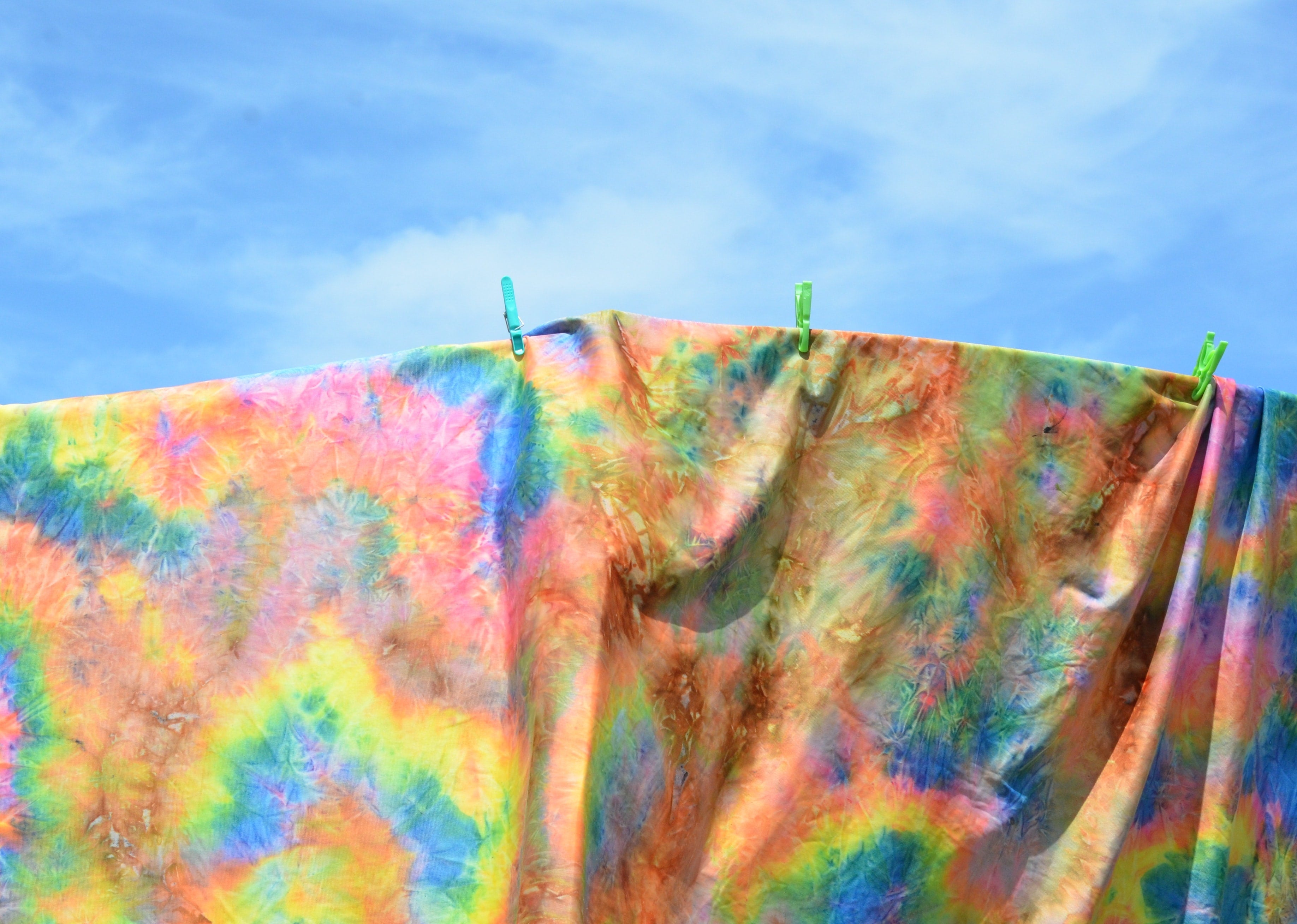 Sheet with colourful tie dye hanging on a clothes line with a blue sky above.