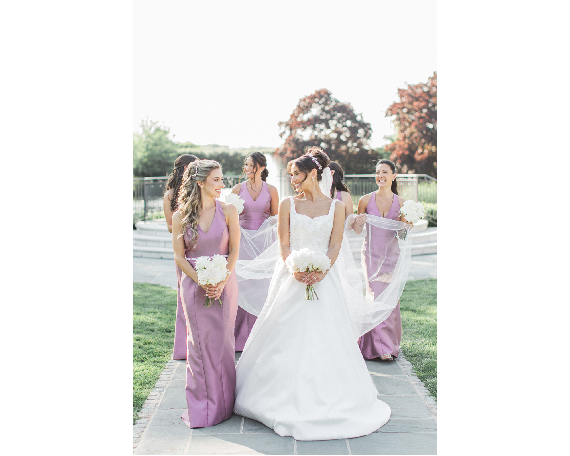 Kara walking outdoors with her bridesmaids, who are wearing lovely orchid dresses. The bride is wearing her Anne Barge gown, a bridal headpiece with Swarovski crystals, freshwater pearls, and porcelain flowers. She's also wearing a crystal-edged veil, and pearl and crystal earrings.