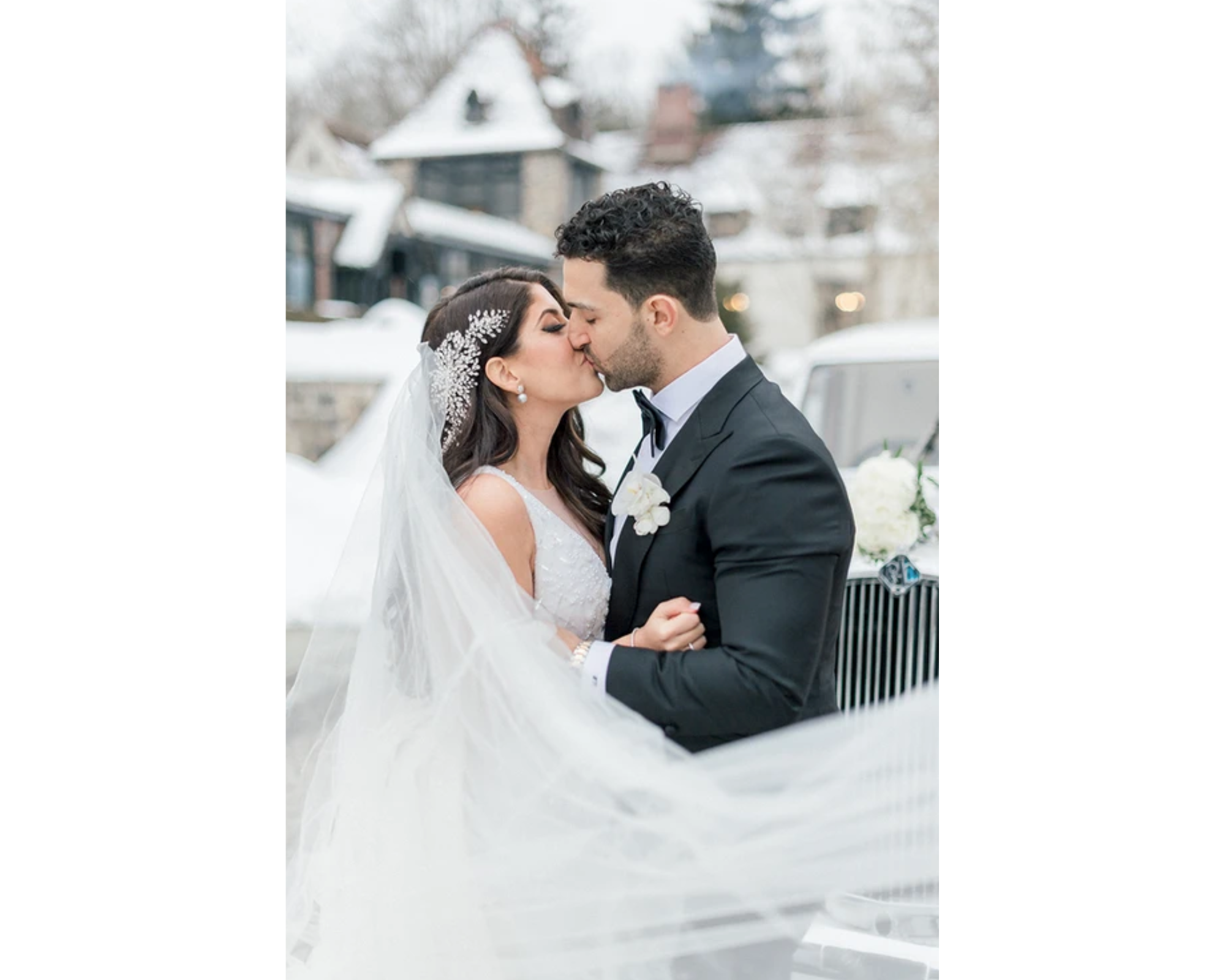 A beautiful bride and groom kiss outside in the snow. Victoria’s cathedral veil flutters in the wind, and her Swarovski crystal headpiece sparkles!