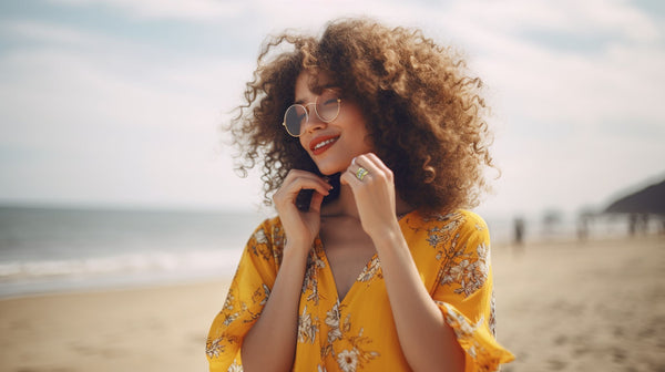 curly haired woman wearing a yellow floral summer dress and showing off her gold cocktail ring on a sunny summer day at the beach