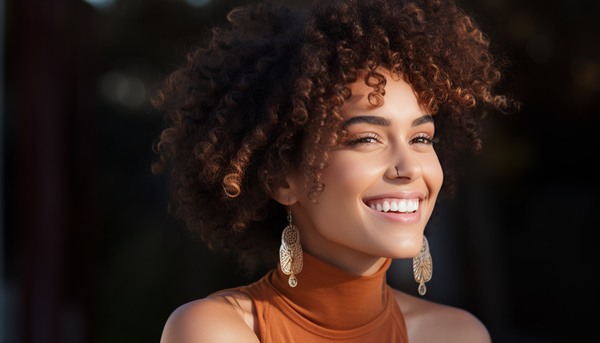 smiling curly haired woman wearing drop earrings, nose ring, and an orange top against dark background