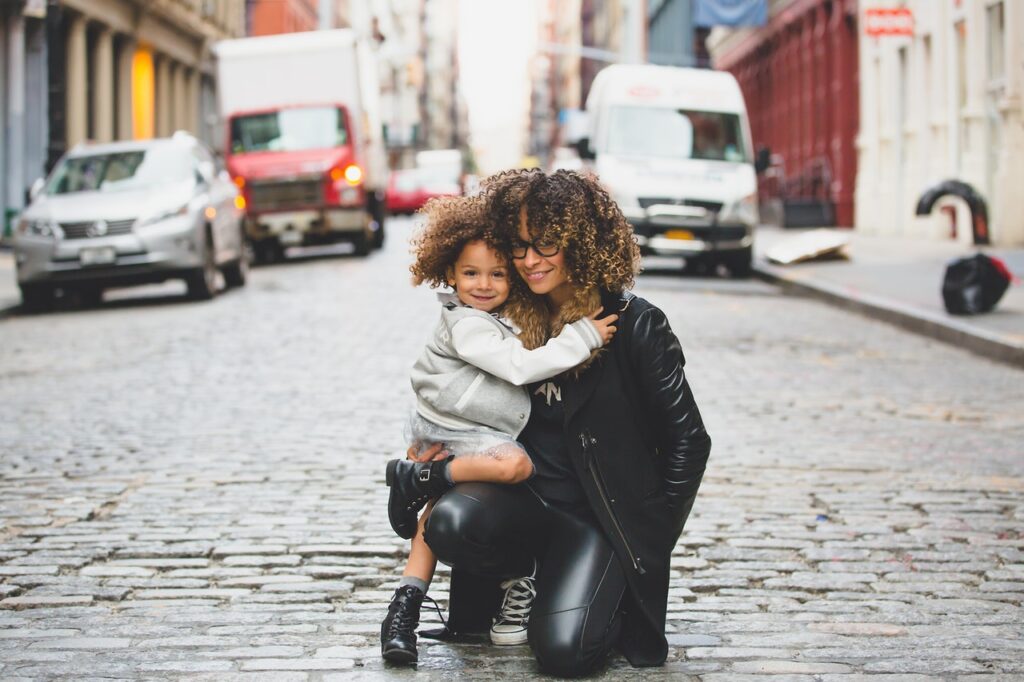 Smiling Curly-Haired Woman Wearing Black Jacket Holding Curly-Haired Girl in the Middle of the Street