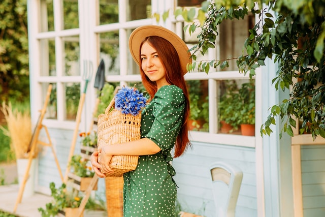 smiling woman in green polka dot dress and hat holding a basket with blue flowers