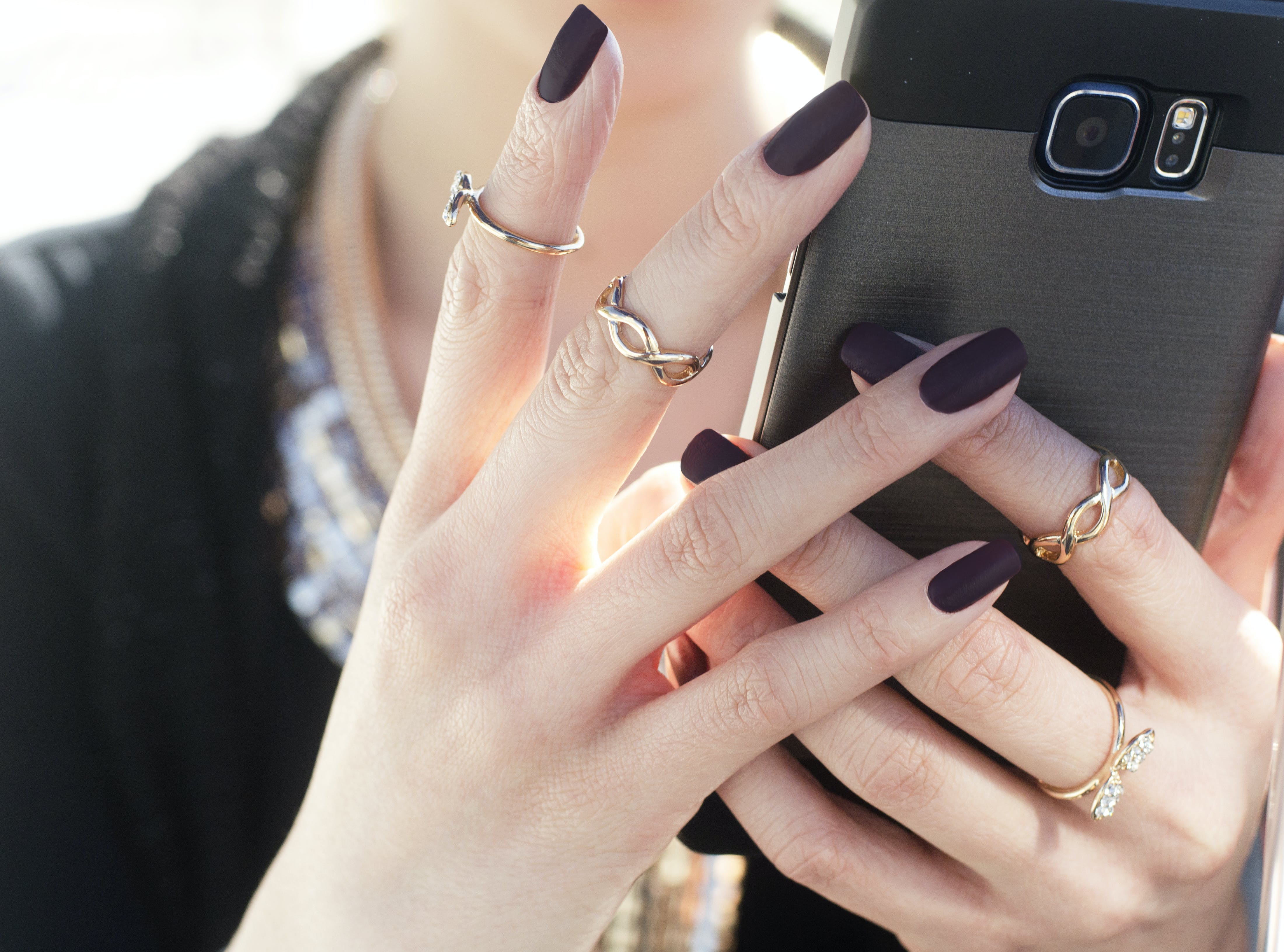 woman with dark plum nails wearing minimalist rose gold rings holding a smartphone