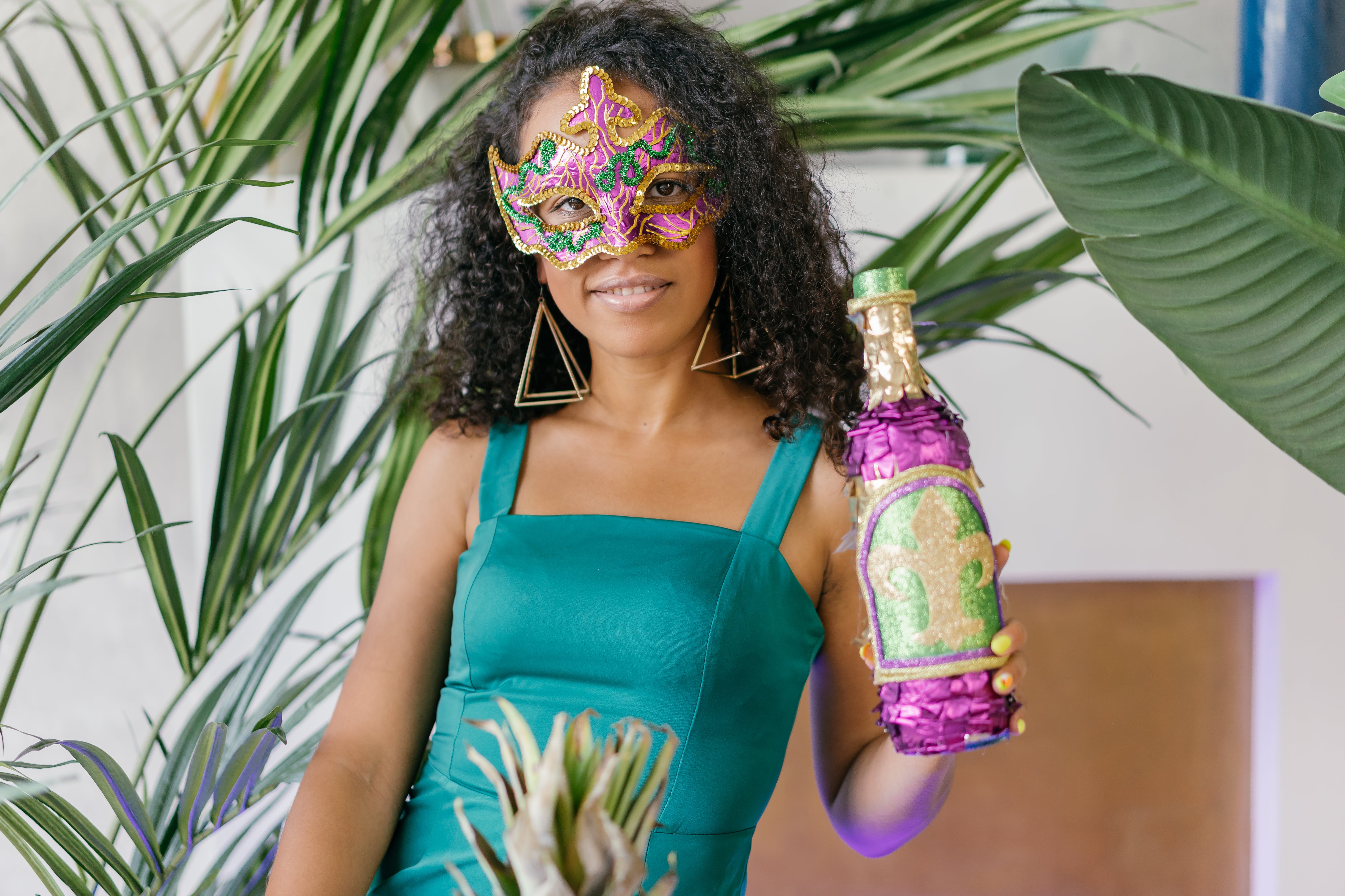 woman wearing green tank top and a mardi gras masquerade mask, holding up a bottle with the dleur de lis symbol