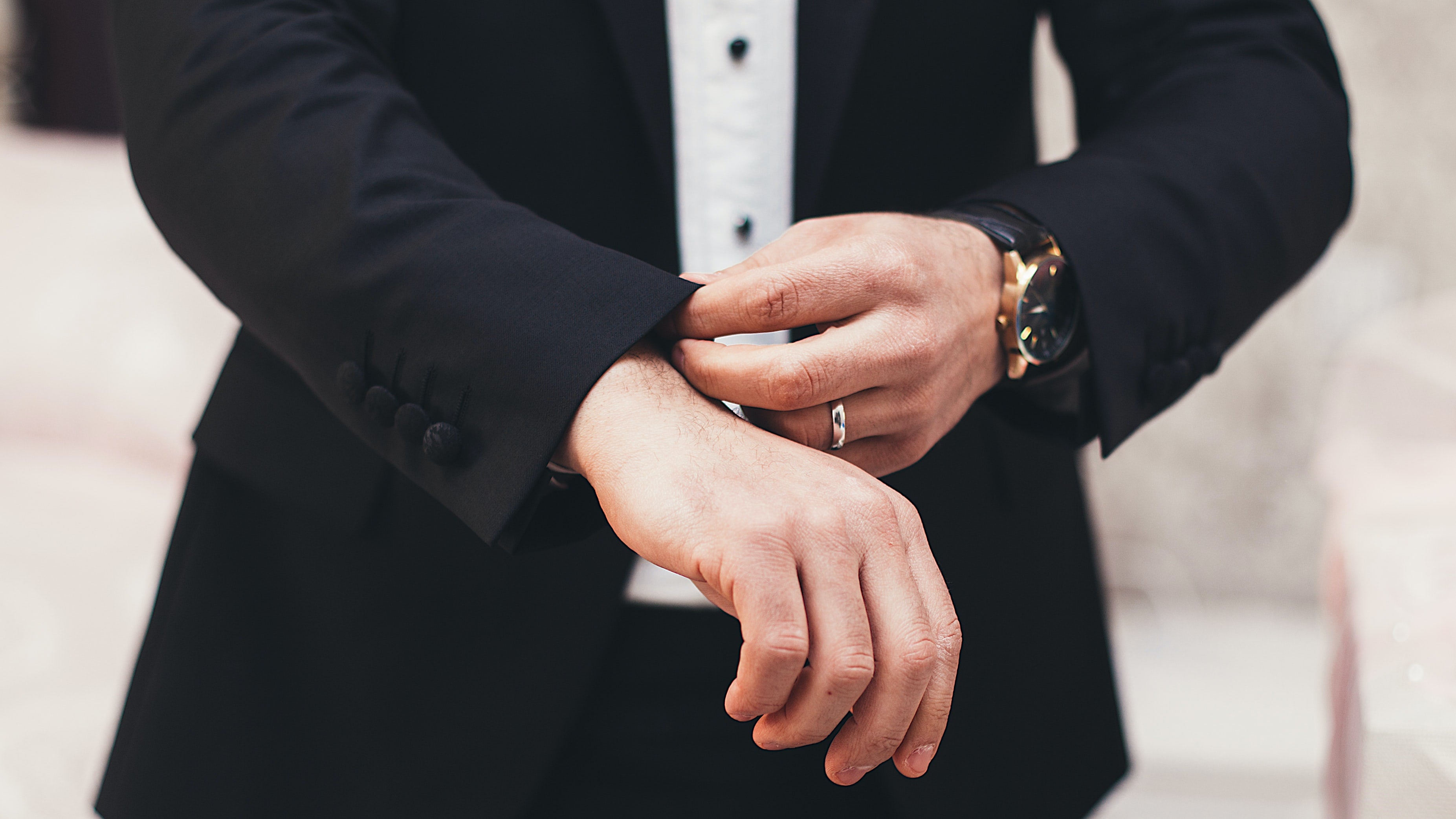 man wearing a silver wedding band and black leather strap watch fixing the sleeves of his black suit