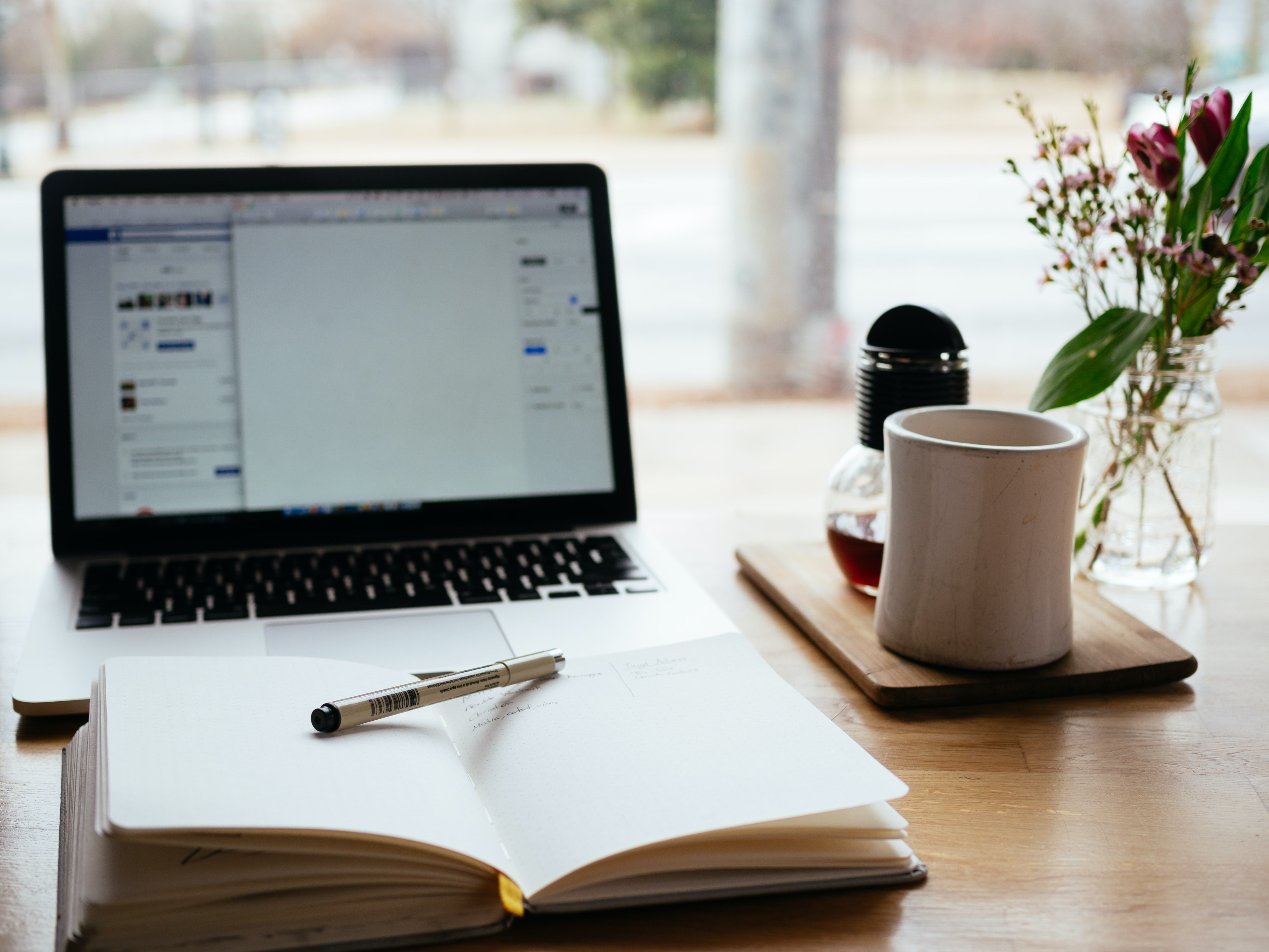 black laptop with Facebook on the screen, an open notebook, a pen, and a mug on a wooden table