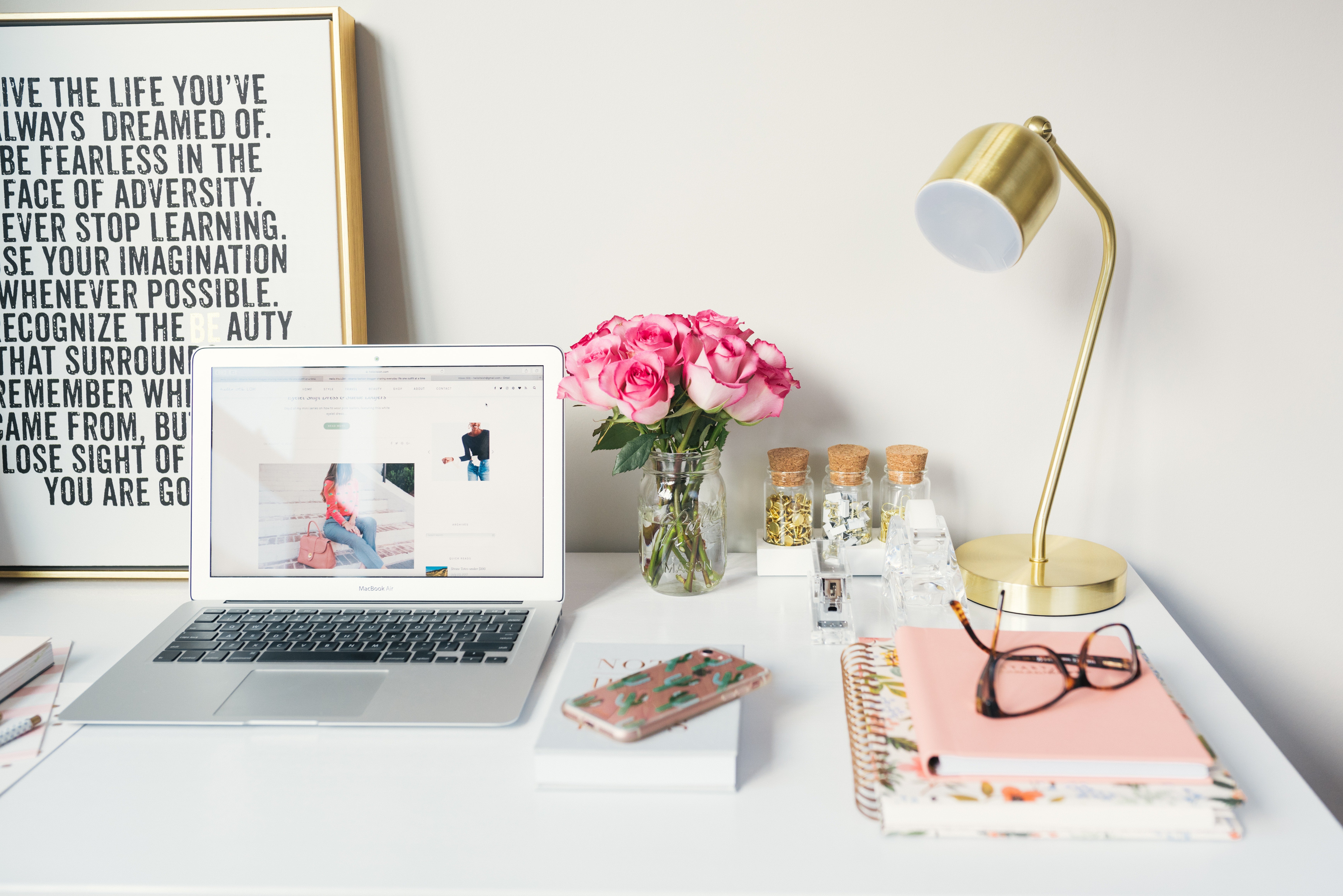 silver laptop with an ecommerce website on the screen, rose gold phone, notebooks, golden table lamp, and pink flowers on a white table