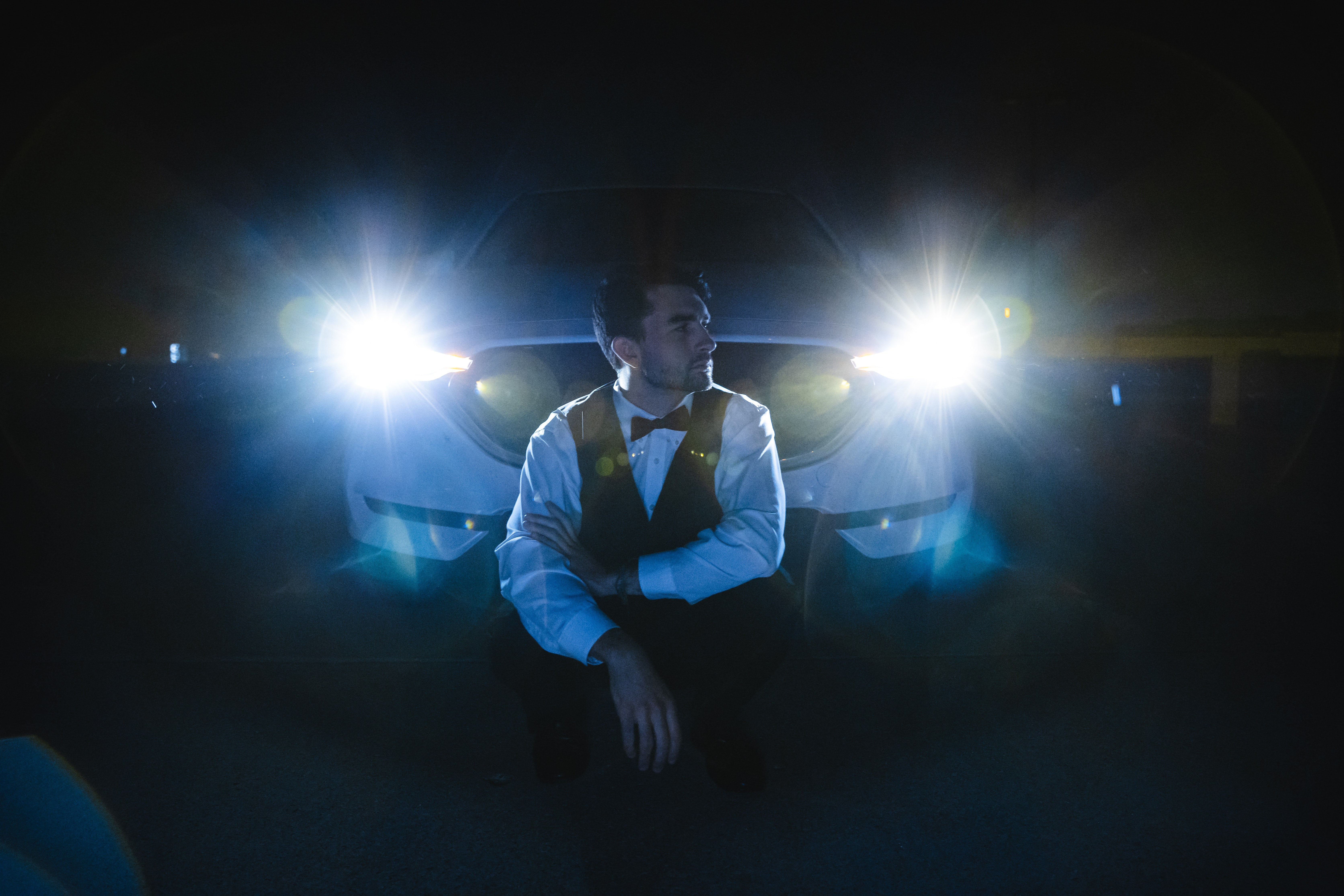 man wearing a black bow tie in front of a white car with its headlights on