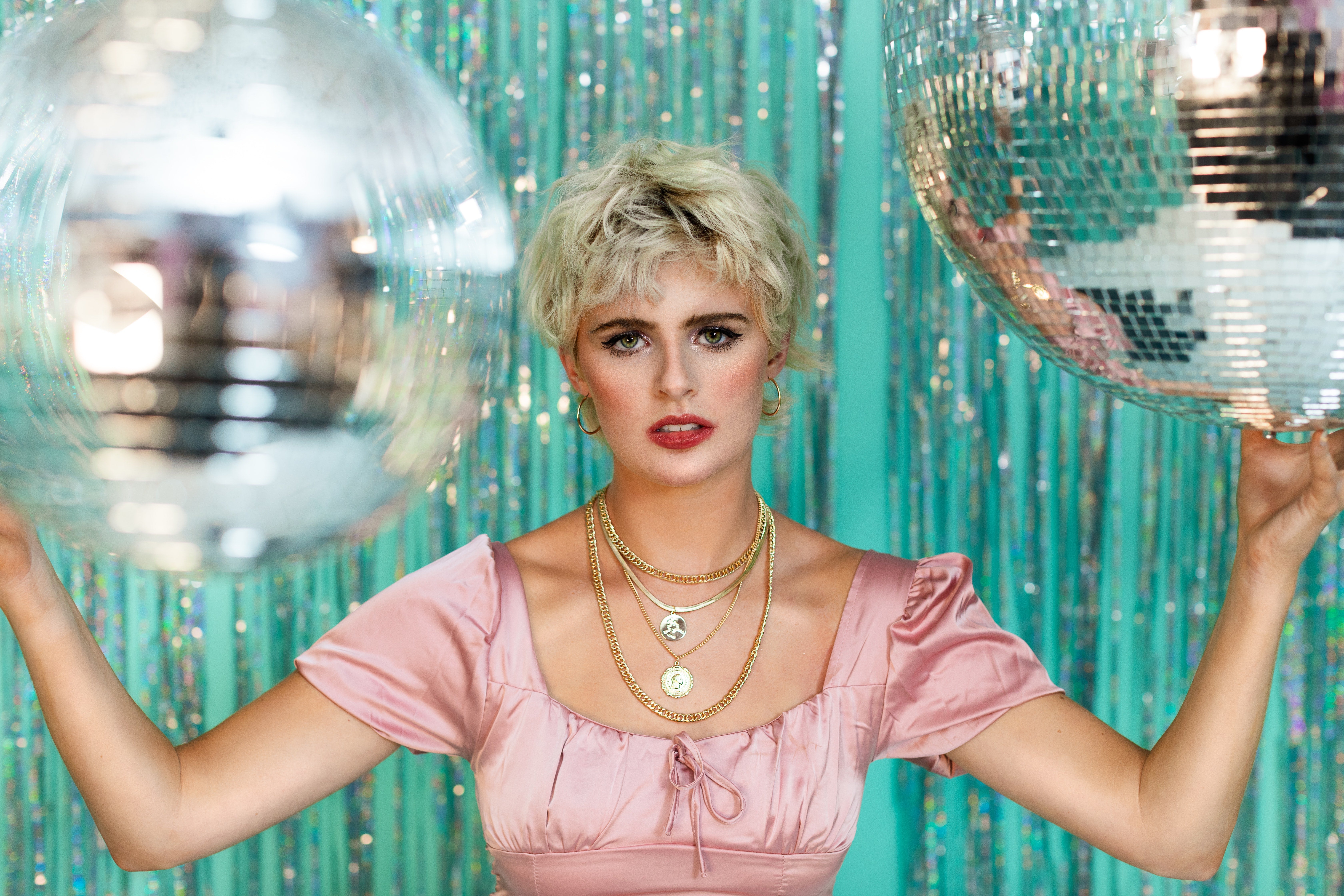 short-haired blonde woman wearing a vintage pink dress and gold necklaces and holding a disco ball on each hand