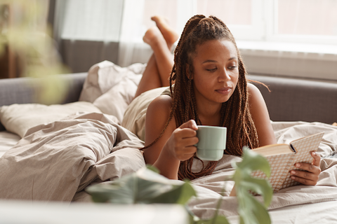 woman lying down reading book in bed holding mug