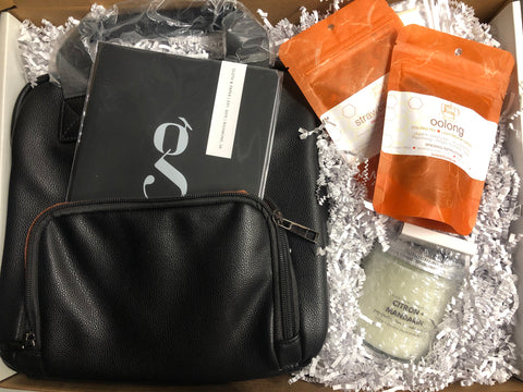 gift box including orange packets of loose leaf tea, a black notebook with a g on it, a candle and a black tote bag