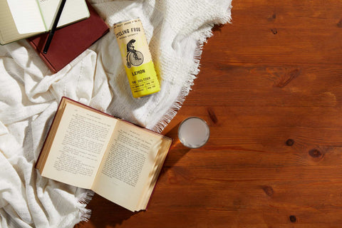 Cycling Frog Lemon Light seltzer on a hardwood floor, next to a book and a cozy blanket.