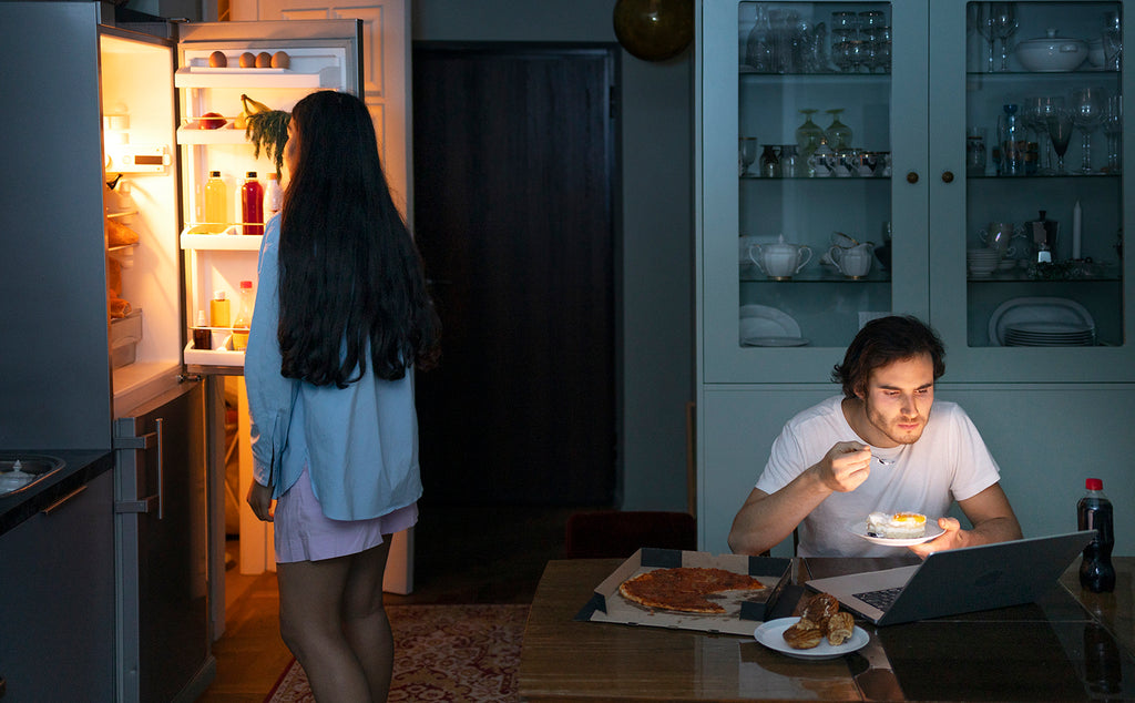 Un hombre mira una computadora y come un pastel, mientras una mujer va al refrigerador para buscar algo para comer.