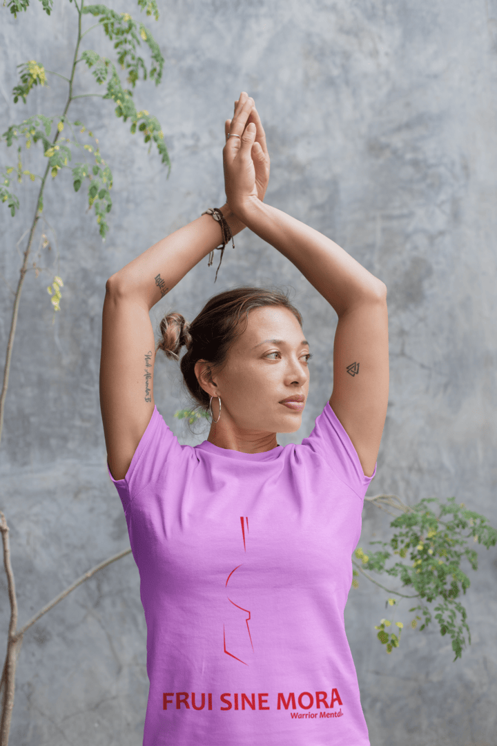 Femme en position yoga, bras au-dessus de la tête, mains jointes : elle porte un t-shirt rose avec un léger dessin représentant une moitié de casque sous la forme d’une ligne rouge. Au bas, est écrit Frui Sine Mora
