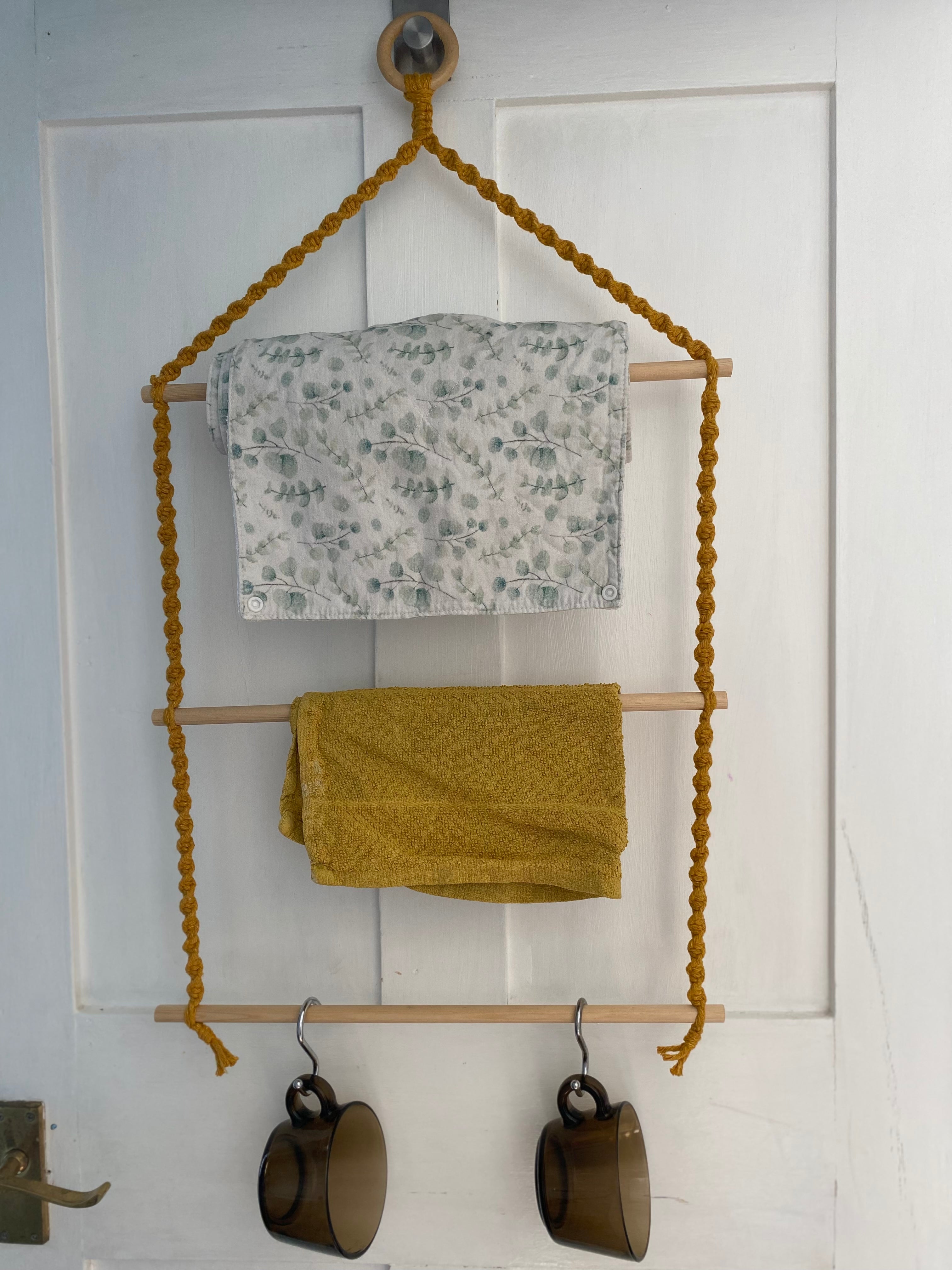 A white wooden door displays a handmade 3 tier wooden kitchen organiser with mustard yellow braided cords. The Macra-Made-With-Love boho farmhouse cottage decor rack has three wooden rods. The top rod holds a recycled cotton white and green patterned towel, the middle rod holds a mustard yellow towel, and the bottom rod holds two brown mugs.