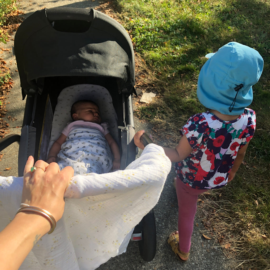 baby our for a walk in stroller with toddler and mums hand on the stroller.  There is a muslin swaddle for shade