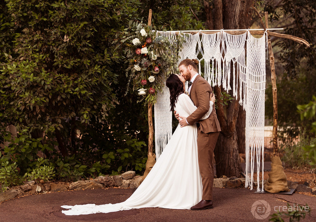Lauren and Alex share a kiss as newly-weds, framed by a bohemian macrame arbour.
