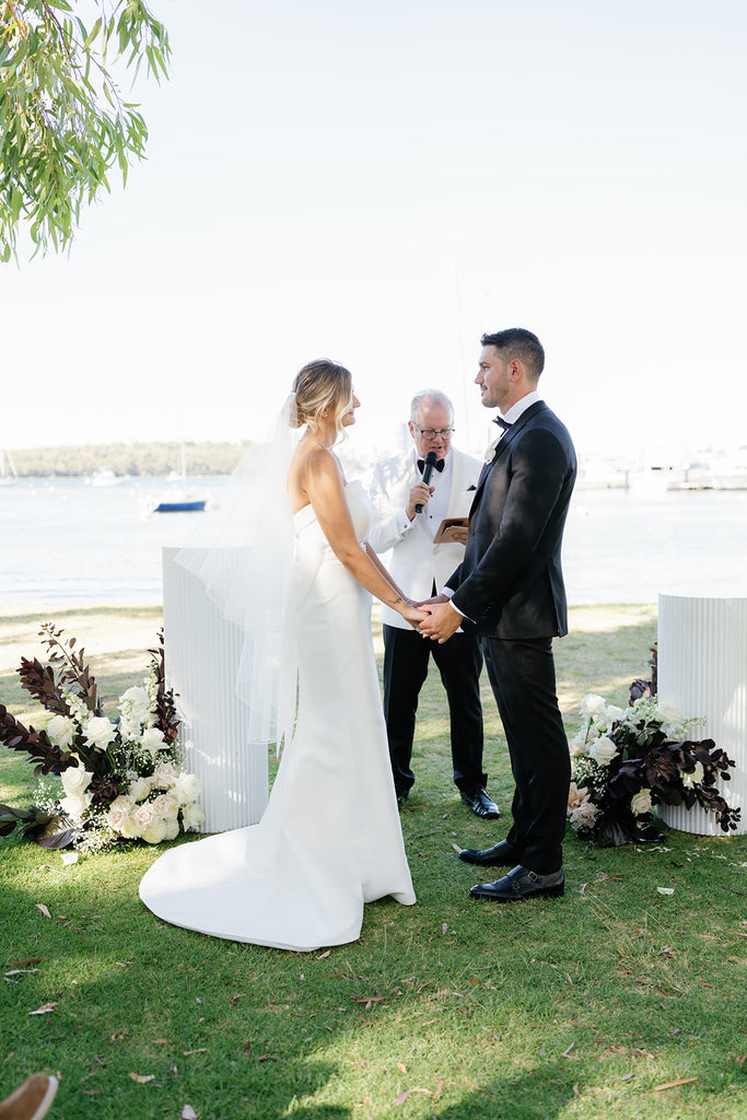 Hannah and Luca hold hands at the end of the aisle; the swan river glistens in the sun behind them at the Matilda Bay foreshore.