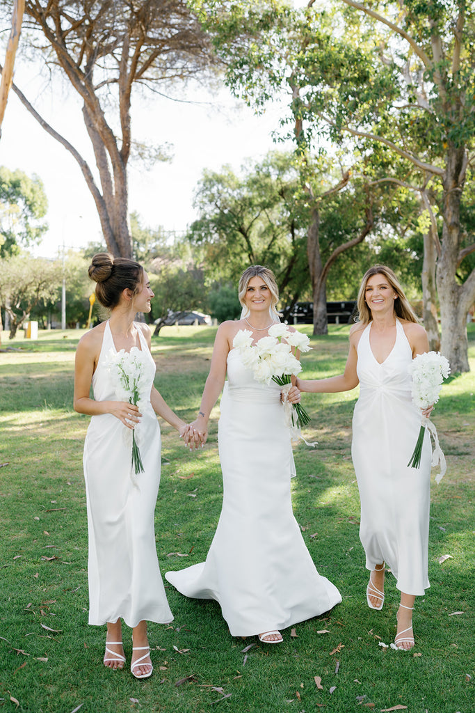 Hannah and her two bridesmaids, dressed in white, walk hand-in-hand.