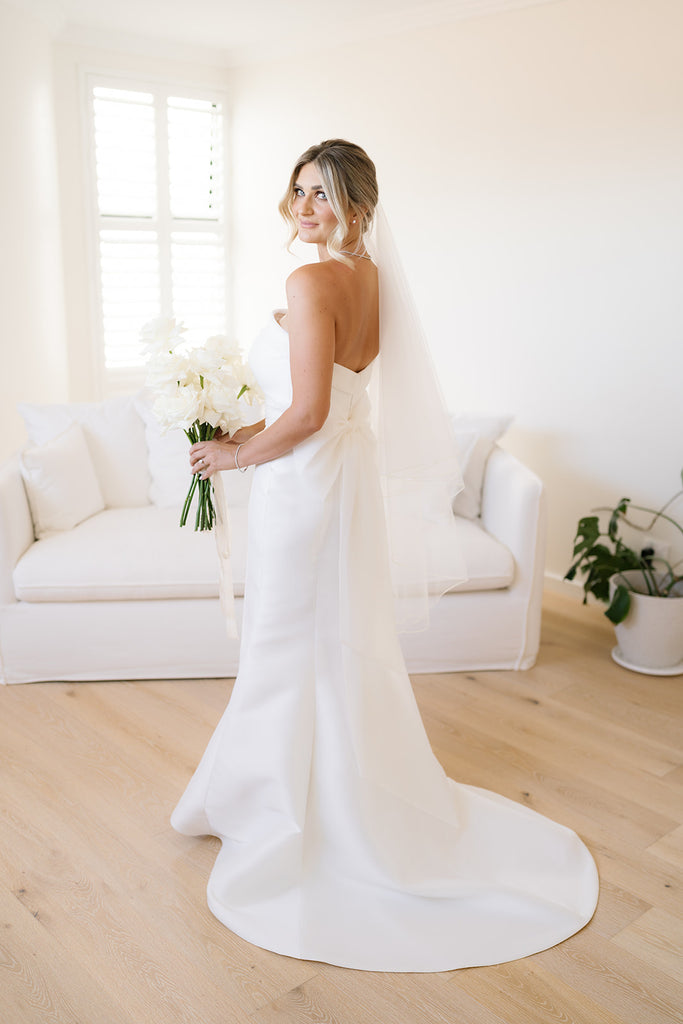 Hannah smiles over her shoulder, dressed in her wedding gown and holding an elegant bouquet of white roses.