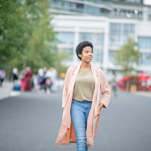 woman with short hair walking wearing a pink trench coats, jeans, and a green top. People walking in the background behind her are blurred.