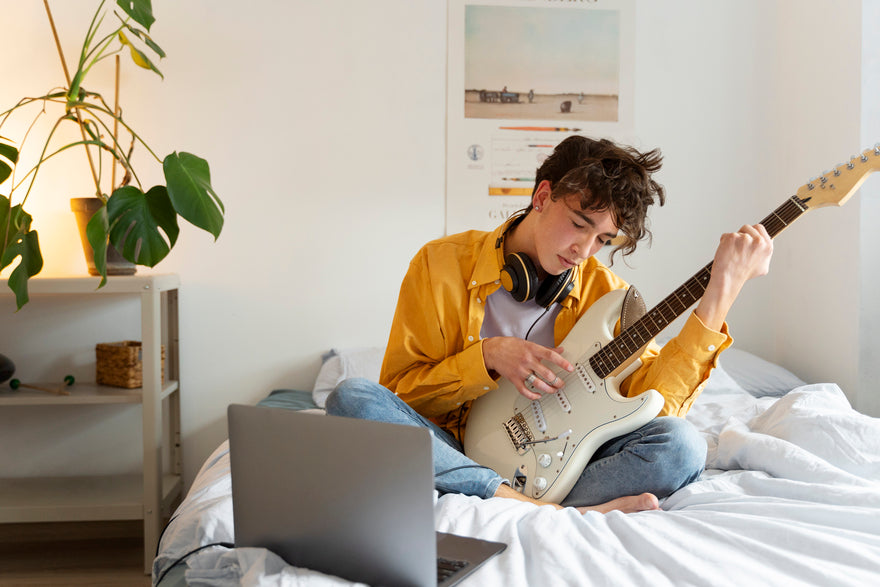 Junge, der auf Bett sitzt und Gitarre hält und auf Laptop schaut