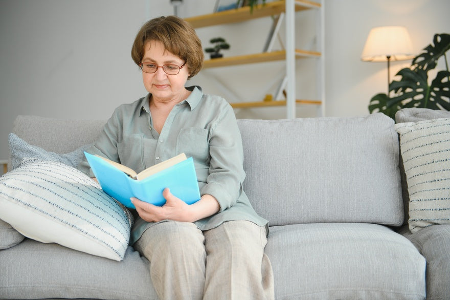 Eine ältere Dame trägt eine Brille und liest ein Buch auf dem Sofa