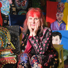 Michelle has dyslexia and is a disabled artist. She has dyed red hair and holds her face in her hands. She looks a little uncertain. Her paintings are all on canvas in the background. They are bright and colourful.