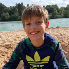 Disabled artist James Tortice smiling into the camera on a sunny beach. James has brown dishevelled hair, a blue jumper with a neon yellow adidas symbol on it.