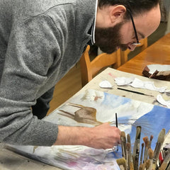 Picture of disabled artist Nick Mair working on a painting, leaning over the canvas. Head down. He has a dark brown hair and beard, wears glasses and a grey jumper.