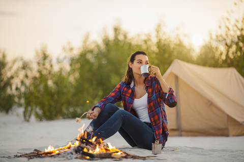 woman in jeans sitting at campsite and drinking hot beverage