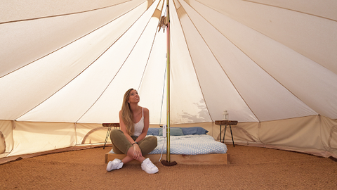 A lady sitting inside her tipi tent on a low bed that is set on a brown cloth